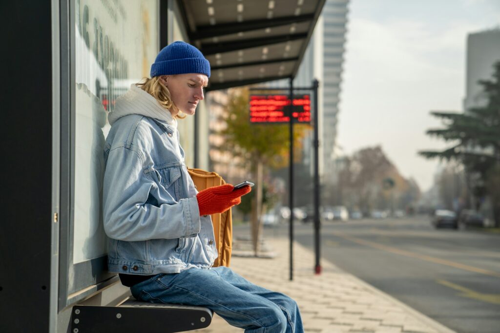 Caucasian millennial guy student with smartphone at bus station in early morning,