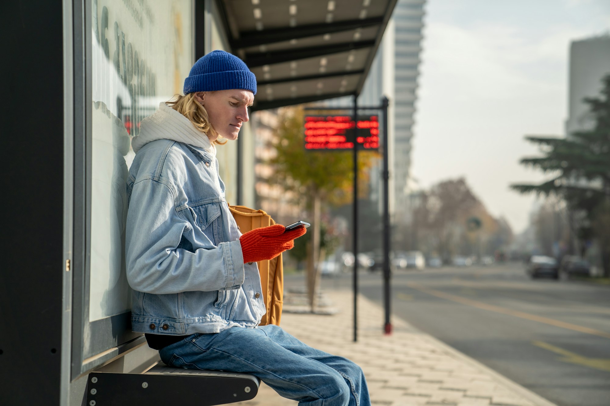 Caucasian millennial guy student with smartphone at bus station in early morning,