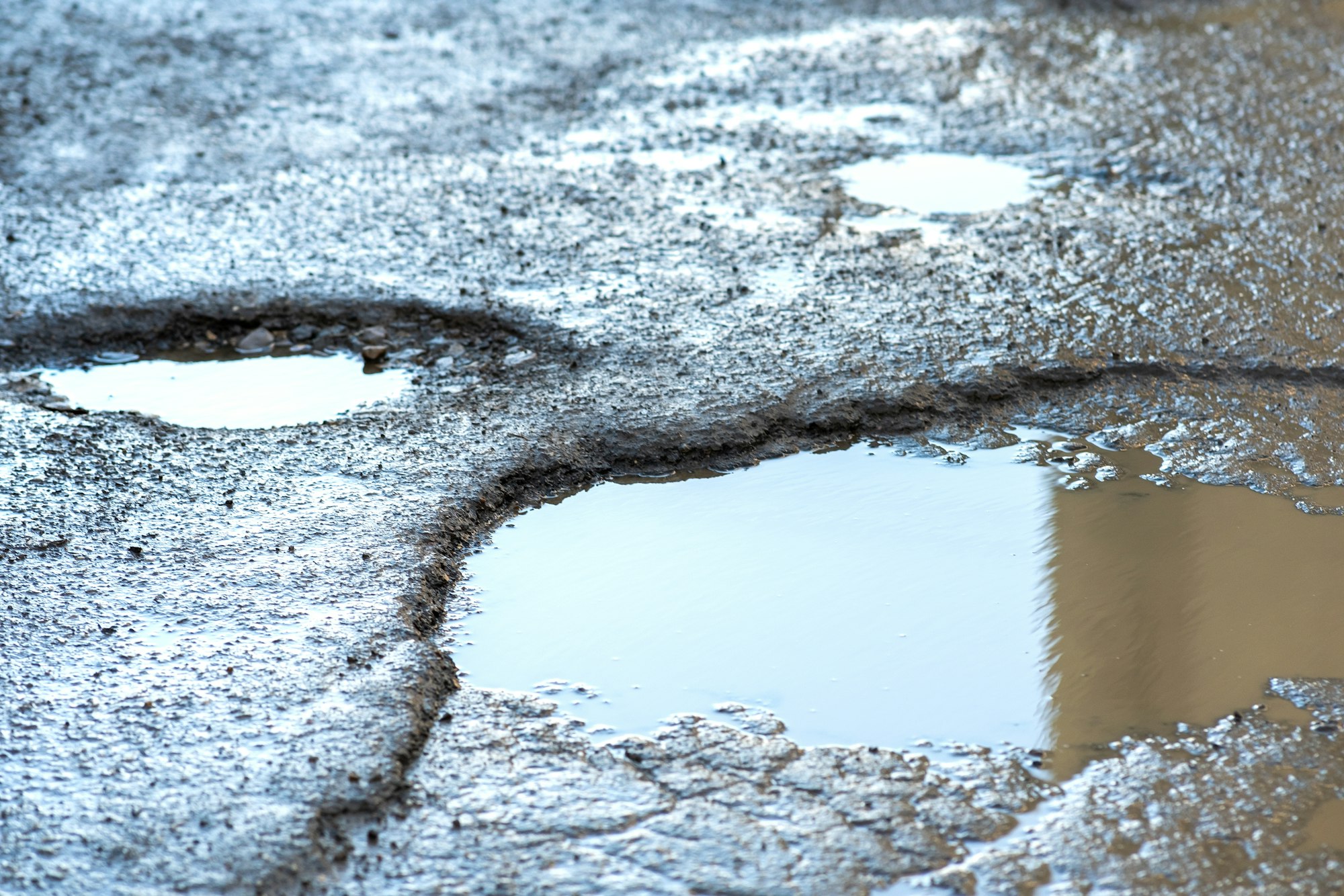 Close up of a road in very bad condition with big potholes full of dirty rain water pools.