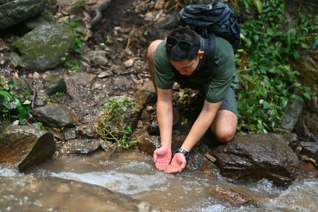 Male tourist with backpack scooping clean water in mountain stream.